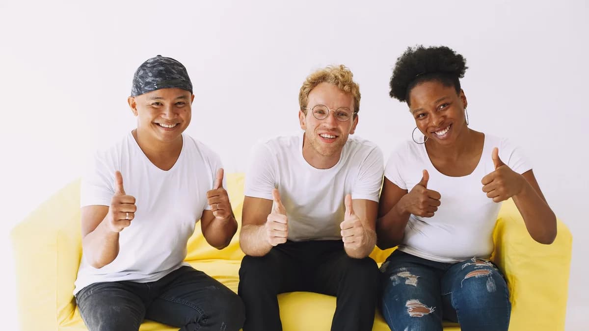 Drei Freunde sitzen auf einem gelben Sofa und zeigen lachend den Daumen nach oben