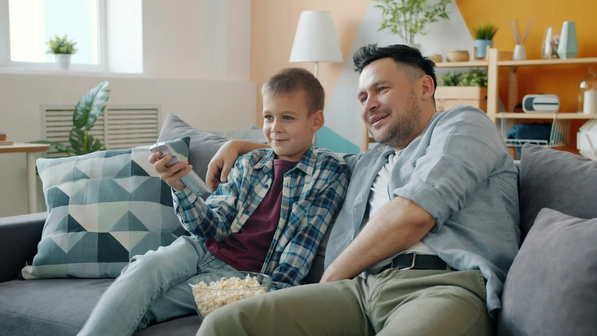 Vater und Sohn sitzen entspannt auf dem Sofa, einer hält eine Fernbedienung, Schüssel mit Popcorn auf dem Tisch