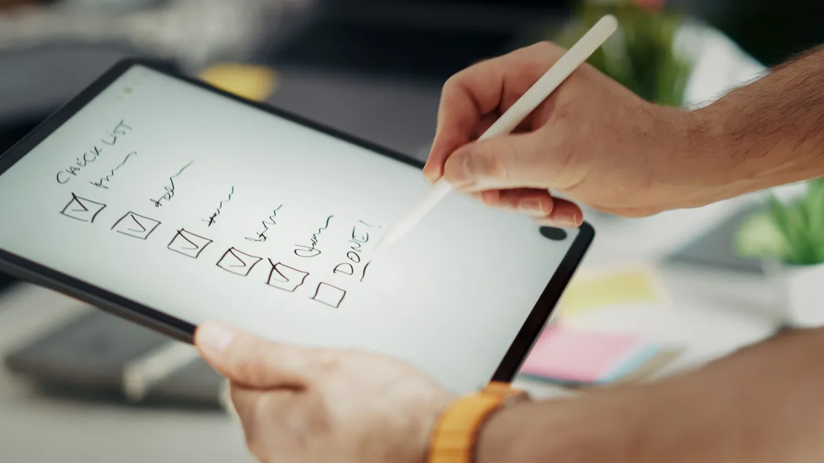 Hand schreibt mit einem Stift auf einem Tablet eine handschriftliche Checkliste mit abgehakten Punkten