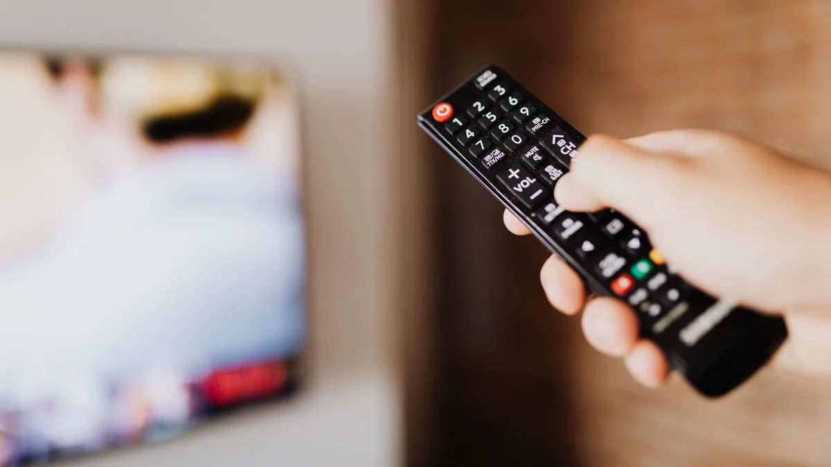 Hand hält eine Fernbedienung und zeigt damit auf einen unscharfen Fernsehbildschirm