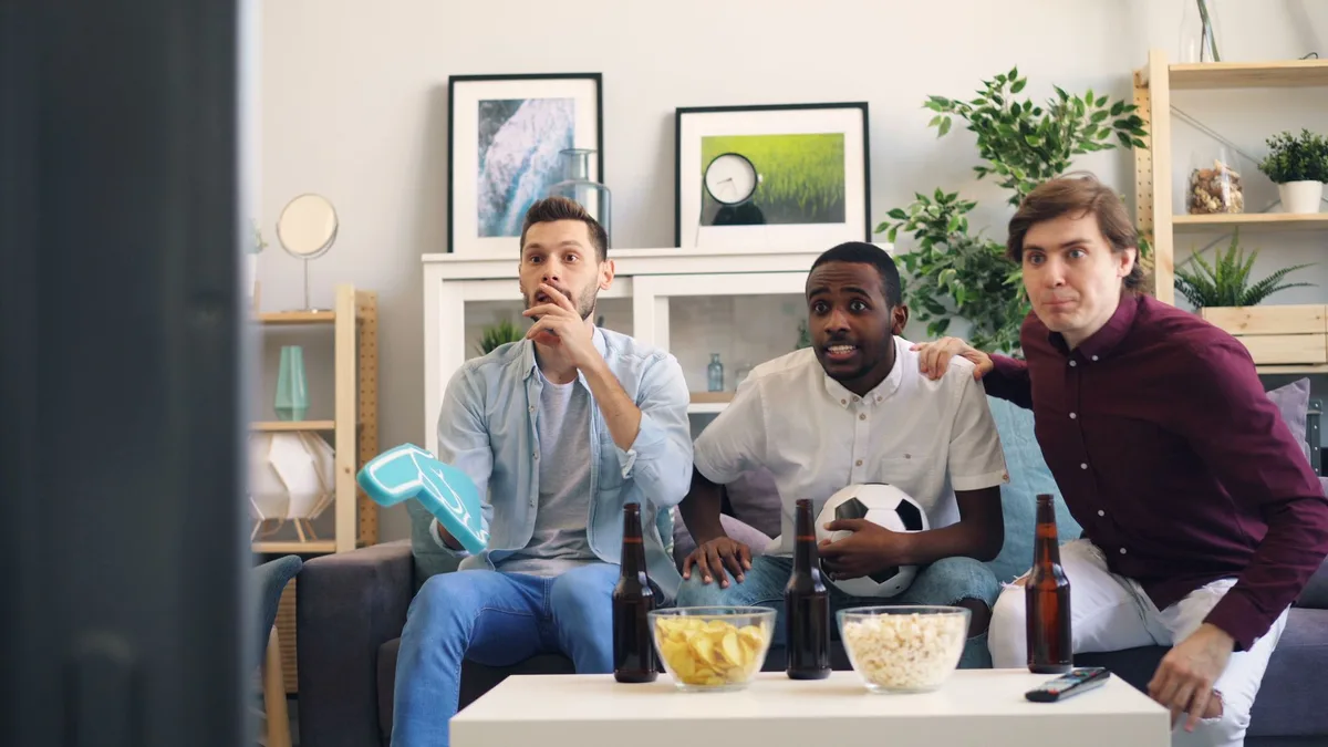 Drei Freunde schauen gemeinsam auf dem Sofa ein Fußballspiel mit Snacks auf dem Tisch